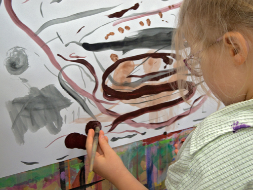 Die Abbildung zeigt ein weiteres Malkind im Malort beim Malen eines Details auf seinem an der Wand aufgespannten Blatt Papier.