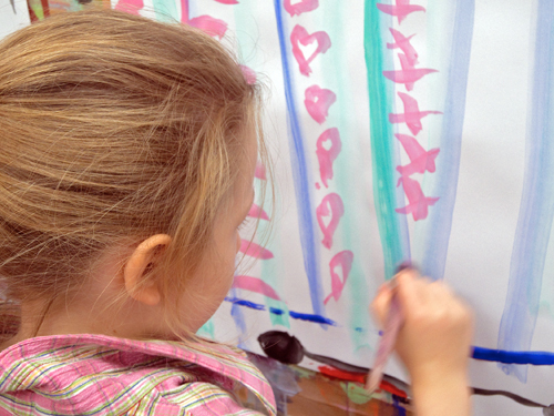Die Abbildung zeigt ein Malkind im Malort beim Malen eines Details auf seinem an der Wand aufgespannten Blatt Papier.